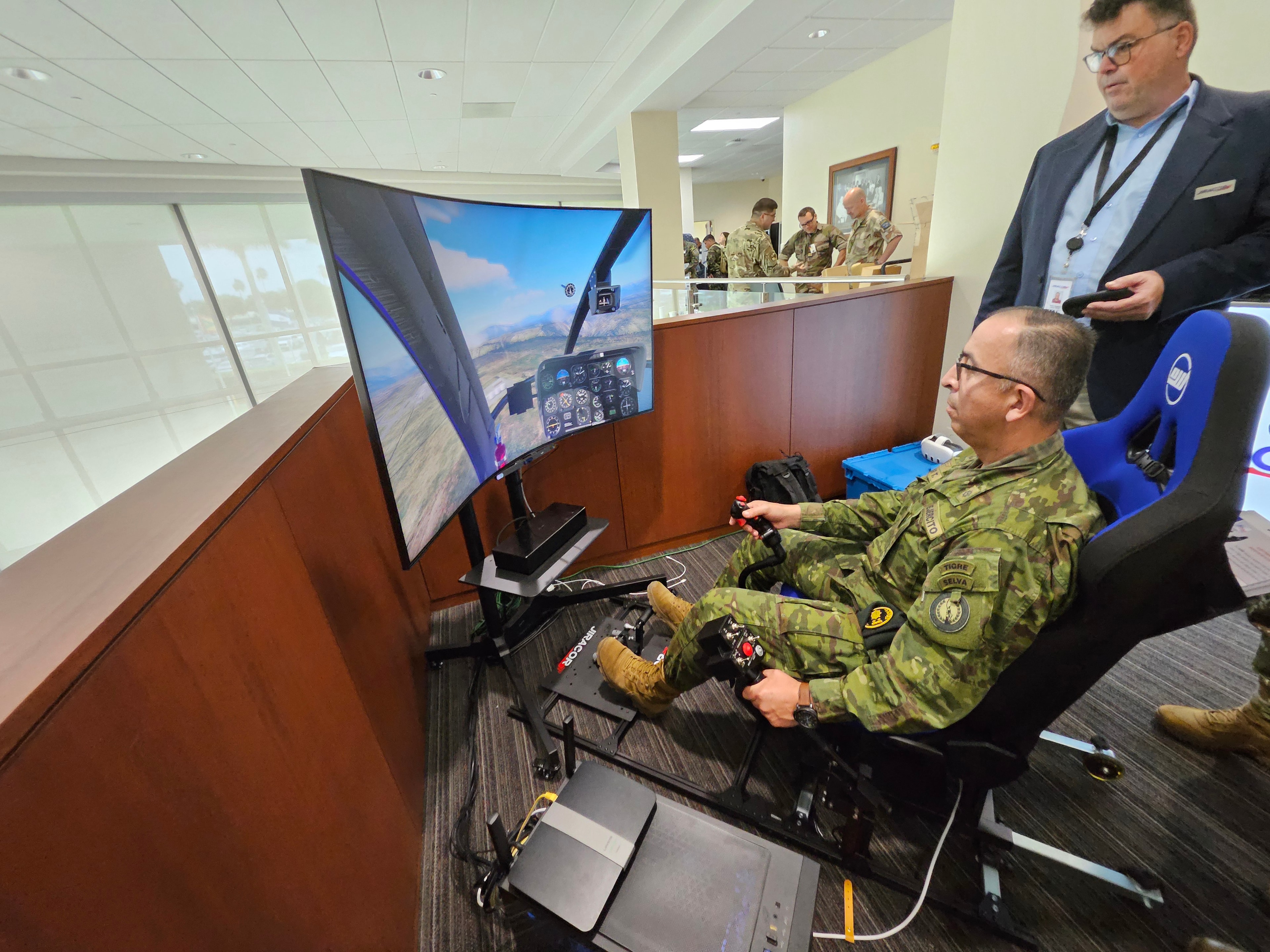 Military representative operating a flight simulator with controls and large curved display during a training technology demonstration.